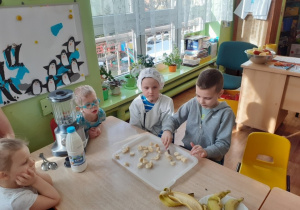 Czworo dzieci siedzi przy stole. Chłopiec kroi banany, reszta dzieci obserwuje.