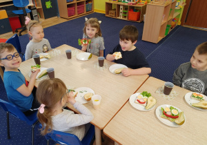 W klasie przy stolikach siedzą dzieci i wykonują kolorowe kanapki z różnych produktów.