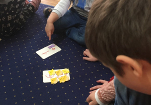 Dzieci siedzą na dywanie przed sobą mają kartki z cyfrą i odpowiednią liczbą elementów.