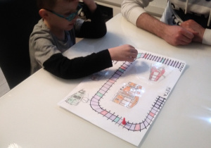 Po lewej stronie siedzi chłopiec i trzyma pionek . Po prawej stronie siedzi mężczyzna i gra w grę planszową. Na środku stołu leży gra planszowa.