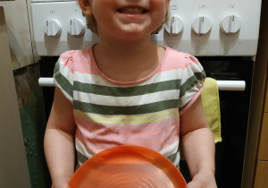 Na zdjęciu widać dziewczynkę, która trzyma pomarańczowy talerz. Na talerzu kanapka z warzywami