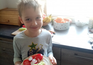 Chłopiec stoi w kuchni i trzyma talerz. Na talerzu kolorowe kanapki z ogórkiem, pomidorem, rzodkiewkami.