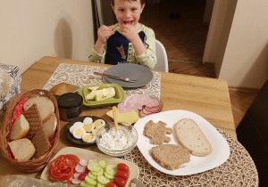 Na środku zdjęcia, przy stole siedzi chłopiec. Na stole kilka talerzy z chlebem, warzywami, sałatą, nabiałem.