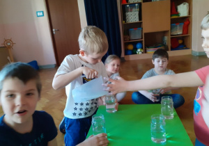 Dzieci siedzą przy stoliku. Jeden chłopiec wlewa płyn do słoika.
