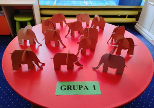 na okrągłym, czerwonym stoliku stoją papierowe słonie wykonane z papieru
