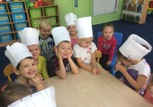 Dzieci w czapkach kucharskich siedzą przy stole i czekają na rozdzielenie zadań.