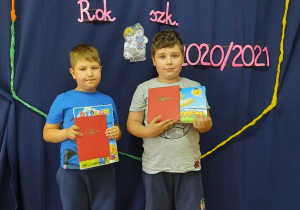 Dwaj chłopcy na tle dekoracji, trzymają w rękach dyplomy i pamiątkową książkę.