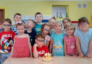 za stolikiem stoi grupa dzieci, na stoliku tort ze świeczką