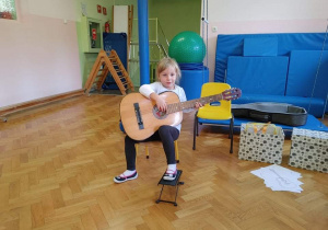 Dziewczynka siedzi na krzesełku, trzyma gitarę