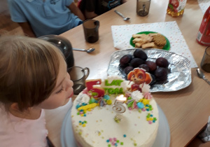 na środku i przodzie widać tort urodzinowy, dziewczynka dmucha świeczki, dzieci siedzą przy stoliku