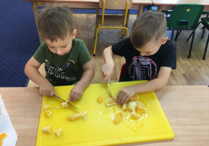 Chłopcy siedzą przy stole i kroją na desce pomarańczę.