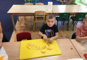 Chłopiec siedzi przy stole i kroi na desce mandarynki.