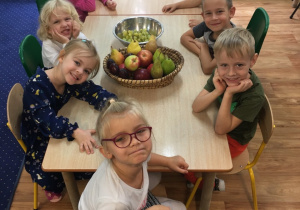 Dzieci siedzą przy stołach na których leżą owoce.