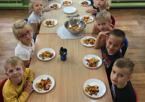 Dzieci siedzą przy stołach, a na nich stoją talerze z sałatką owocową wykonaną przez dzieci.