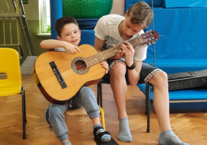 Chłopiec siedzi na krzesełku i próbuje grać na gitarze z pomocą starszego kolegi.