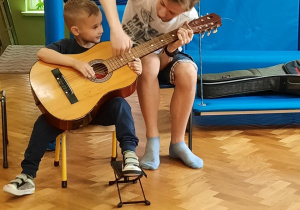 Chłopiec siedzi na krzesełku i próbuje grać na gitarze z pomocą starszego kolegi.