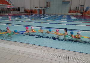 Dzieci w basenie płyną jeden za drugim.
