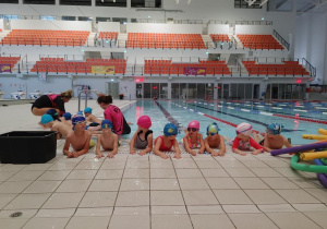 Dzieci w basenie wykonują ćwiczenia przygotowujące je do nauki pływania.