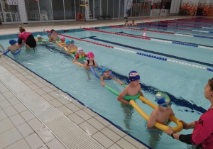 Dzieci w basenie wykonują ćwiczenia przygotowujące je do nauki pływania.