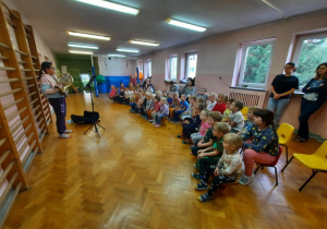 Przedszkolaki siedzą na sali gimnastycznej i słuchają koncertu w wykonaniu uczennicy szkoły muzycznej, która gra na waltorni -rogu.