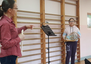 Po lewej stoi pani od rytmiki i trzyma w dłoni flet prosty. Z prawej uczennica szkoły muzycznej trzyma waltornię.
