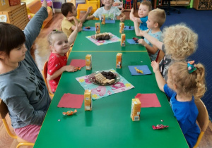 Dzieci siedzą przy stole na którym stoją słodkości i wskazują na jubilata.