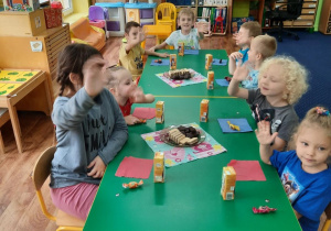 Dzieci siedzą przy stole zastawionym słodkościami i machają.