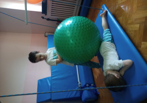 na dużej huśtawce leży chłopiec, na jego plecach duża piłka, którą trzyma chłopiec