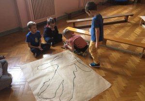 czterech chłopców klęczy na podłodze, przed nimi duży karton, na którym odrysowana jest postać dziecka