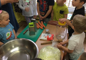 Dzieci zgromadzone wokół stołu na którym leżą produkty i materiały niezbędne do kiszenia kapusty. Pani prezentuje główkę kapusty.