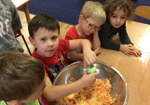 Dzieci siedzą przy stole. Chłopiec dokłada przyprawy do miski z poszatkowaną kapustą i marchewką.
