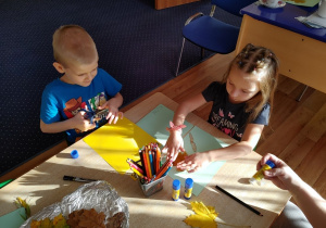 Dzieci siedzą przy stoliku i naklejają materiał przyrodniczy na kartki.