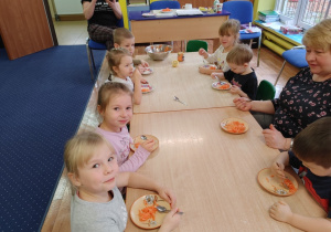 Dzieci siedzą przy stołach i zjadają samodzielnie przygotowaną surówkę marchewkową.