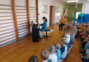 Dziewczynka gra na pianinie, a przedszkolaki słuchają.