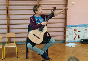 Uczeń prezentuje sprzęt gitarzysty siedząc na krześle.