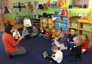 Dzieci z nauczycielem siedzą w kole na dywanie i słuchają go.
