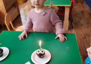 Dziewczynka stoi przy stole przed babeczką z zapaloną świeczką.