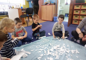 Dzieci siedzą na podłodze wokół materiału i drą papier.