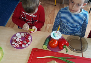 Dzieci siedzą przy stole i kroją produkty z których przygotują kanapki.