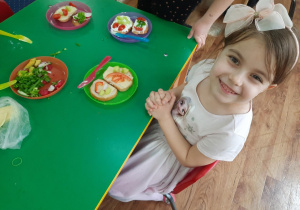 Dziewczynka siedzi przy stole na którym stoi talerz z jej kanapką, którą wykonała samodzielnie.