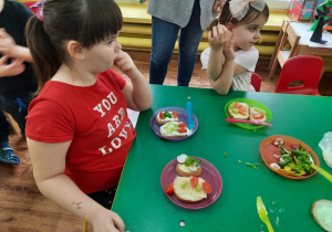 Dziewczynki siedzą przy stole na którym stoją talerze z kanapkami, którą wykonały samodzielnie.