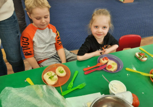 Dzieci siedzą przy stole na którym stoją talerze z kanapkami, które wykonały samodzielnie.