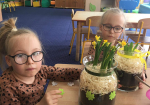 dwie dziewczynki siedzą przy stoliku, na nim ozdobne słoje z kwiatami
