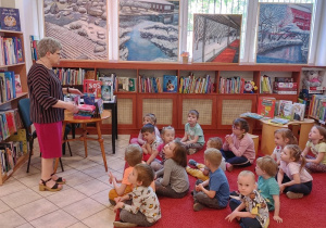 dzieci siedzą na czerwonym dywanie w bibliotece, pani bibliotekarka pokazuje książki