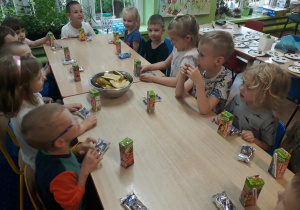 Dzieci siedzą przy stole na którym stoją soczki , ciasteczka i owoce.
