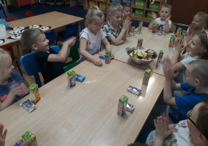 Dzieci siedzą przy stole na którym stoją soczki , ciasteczka i owoce.