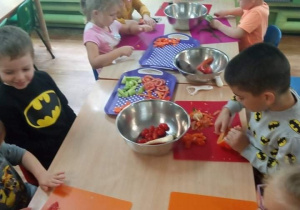 Dzieci siedzą przy stole i kroją warzywa.