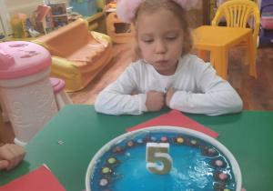 Dziewczynka siedzi przy stole i zdmuchuje świeczkę urodzinową.
