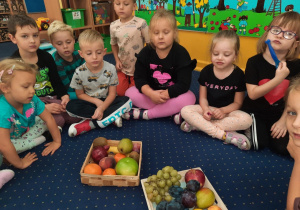 Dzieci siedzą na podłodze w klasie, a przed nimi leżą zakupione owoce.