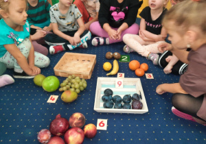 Dzieci siedzą na podłodze w klasie i przeliczają owoce.